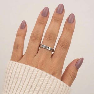 Puede incluir: Un anillo plateado con la fecha grabada "2.12.25" y s&iacute;mbolos de coraz&oacute;n. El anillo se lleva en un dedo con esmalte de u&ntilde;as color malva. La persona lleva un jersey blanco.