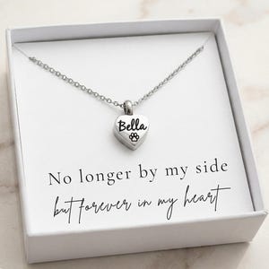 Puede incluir: Un collar con colgante de plata en forma de corazón con el nombre "Bella" y una huella de pata. El collar se muestra en una caja blanca con el texto "No longer by my side but forever in my heart."