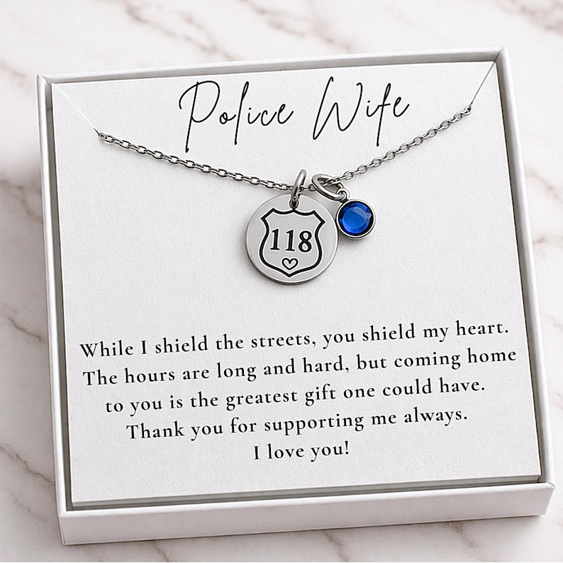 K&ouml;nnte beinhalten: Eine silberne Halskette mit einem Anh&auml;nger "Police Wife". Der Anh&auml;nger hat ein Abzeichen mit der Nummer "118" und einen blauen Edelstein. Die Karte lautet "While I shield the streets, you shield my heart."