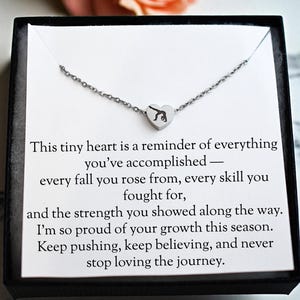 May include: A silver-toned necklace with a heart-shaped pendant featuring a gymnast silhouette. The necklace is presented in a black gift box with a white card that has inspirational text.