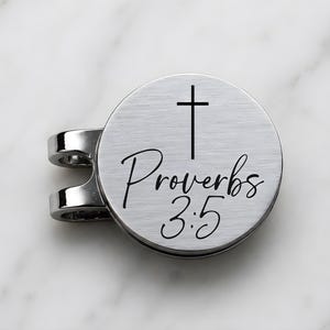 May include: A circular silver-toned metal golf ball marker with a brushed finish. The surface features a black engraved cross symbol positioned above the text Proverbs 3:5 in a cursive script font. The marker has a U-shaped clip attachment.