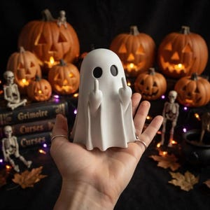 Peut inclure: Une figurine de fantôme blanche avec deux yeux ovales noirs, représentée avec les deux bras levés et les majeurs tendus. Le fantôme au fini mat et lisse est tenu dans une paume sur un fond sombre avec plusieurs citrouilles illuminées et squelettes.