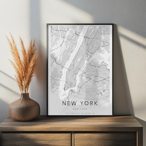Op de afbeelding: Een ingelijste verticale wandkunstprint met een zwart-wit stratenplan van New York City. De print bevat de tekst NEW YORK, NEW YORK en de coördinaten 40.7128 N / 74.0060 W in een strak, schreefloos lettertype in het midden onderaan.