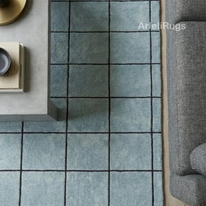 Könnte beinhalten: Ein hellblauer Teppich mit einem dunklen Gittermuster aus sich kreuzenden schwarzen Linien. Der Teppich hat eine strukturierte Oberfläche und enthält den Schriftzug ArieliRugs in Weiß auf der oberen rechten Seite des blauen Stoffes.