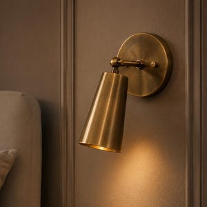May include: A wall-mounted lamp featuring a circular brass backplate and a conical brass shade. The fixture includes an adjustable swivel joint connecting the arm to the shade, which directs light downward, casting a warm glow on the wall surface.