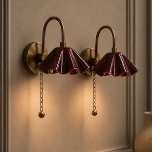 May include: Two wall-mounted lamps featuring curved brass arms and circular backplates. Each lamp has a deep red, glossy, ruffled ceramic shade and a brass pull chain with a small spherical weight, emitting a warm glow from beneath the shade.