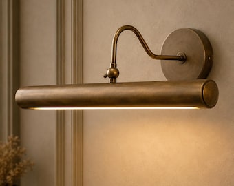 Op de afbeelding: Een wandlamp voor schilderijen met een antieke messing afwerking. Het armatuur is voorzien van een ronde basisplaat, een gebogen arm met een verstelbaar gewricht en een lange, cilindrische horizontale kap die warm licht naar beneden werpt.