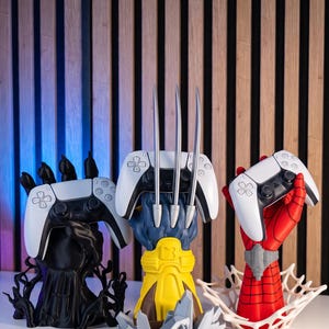 May include: Three controller stands shaped like hands. The left is a black hand with tendrils. The center is a blue and yellow hand with three silver claws. The right is a red and blue hand with a white web base. Each holds a white game controller.