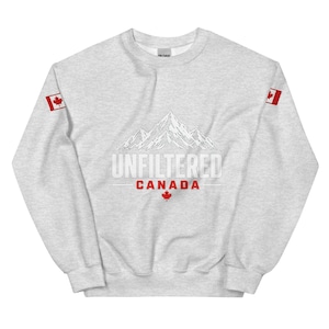 Puede incluir: Una sudadera gris claro de cuello redondo con un gráfico de líneas blancas de montañas en el pecho. Debajo de las montañas, el texto dice UNFILTERED CANADA en blanco y rojo, con una pequeña hoja de arce roja. Cada manga muestra una bandera canadiense.