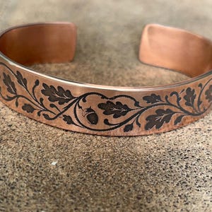 Può includere: Un bracciale a fascia in rame con un motivo scuro inciso raffigurante foglie di quercia e una ghianda. Il metallo ha una finitura liscia e lucida e il disegno si avvolge orizzontalmente attorno all'esterno del bracciale aperto.