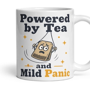 May include: A white ceramic mug featuring the dark blue text "Powered by Tea" and "and Mild Panic" in orange. Between the text is a cartoon illustration of a tan tea bag with a worried facial expression, surrounded by small orange stars.