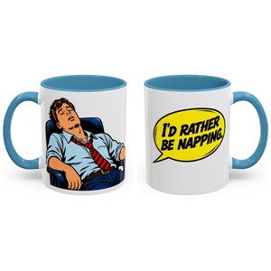 May include: A white ceramic mug with a blue handle and rim. One side features a comic-style illustration of a person sleeping in a chair, wearing a light blue shirt and red striped tie. The other side displays a yellow speech bubble with the text I'D RATHER BE NAPPING.