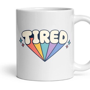 May include: A white ceramic mug featuring the word TIRED in large, bold, cream-coloured letters. Below the text is a colourful, fan-shaped graphic with stripes in red, yellow, teal, purple, and blue. Small four-pointed star icons flank the design.