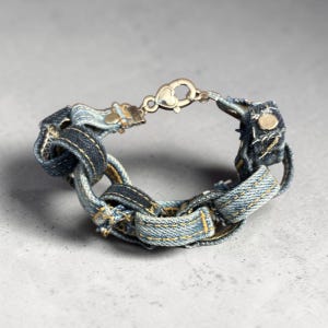 May include: A bracelet constructed from interconnected loops of blue denim fabric with visible yellow stitching. The denim links are joined by a metallic lobster claw clasp featuring a heart-shaped detail, creating a textured, chain-like appearance.
