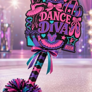 May include: A handheld sign featuring the text DANCE DIVA in purple and pink, decorated with a ballerina, crown, duffel bag, ballet slippers, and a trophy. The handle is wrapped in black ribbon with pink and teal accents and a pom-pom base.
