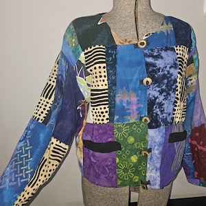 May include: A long-sleeved jacket featuring a patchwork design with various patterns and colors including blue, purple, green, and cream. The garment has a round neckline, five wooden toggle buttons down the front, and two black horizontal pockets.