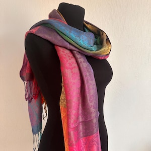 Könnte beinhalten: Ein rechteckiger Schal mit einem lebendigen Farbverlauf in Magenta, Violett, Blau und Gold. Der Stoff zeigt durchgehend ein komplexes, sich wiederholendes Paisley-Muster und ist an den Enden mit zarten Fransen versehen.