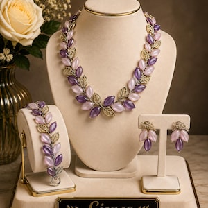 May include: A jewelry set featuring a necklace, bracelet, and earrings with marquise-shaped light pink and deep purple stones interspersed with gold-toned metal leaves. A black plaque at the base displays the gold text Lisner with leaf accents.