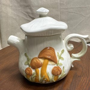 Może przedstawiać: Biały ceramiczny czajniczek z pokrywką w kształcie kapelusza grzyba. Korpus czajniczka ozdobiony jest wypukłymi, trójwymiarowymi ilustracjami brązowych i żółtych grzybów w otoczeniu zielonych liści, posiada zakrzywiony uchwyt oraz dzióbek.