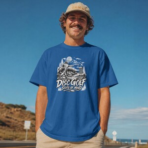 May include: A royal blue short-sleeve t-shirt featuring a white graphic print of a person throwing a disc toward a basket in a mountainous landscape. The text "DISC GOLF STATE OF MIND" is printed in white below the illustration on the chest.