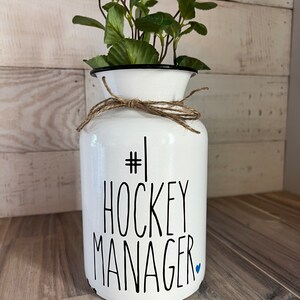 May include: White ceramic vase with a black rim and a brown twine bow. The vase has the text "#1 HOCKEY MANAGER" written in black with a small blue heart.