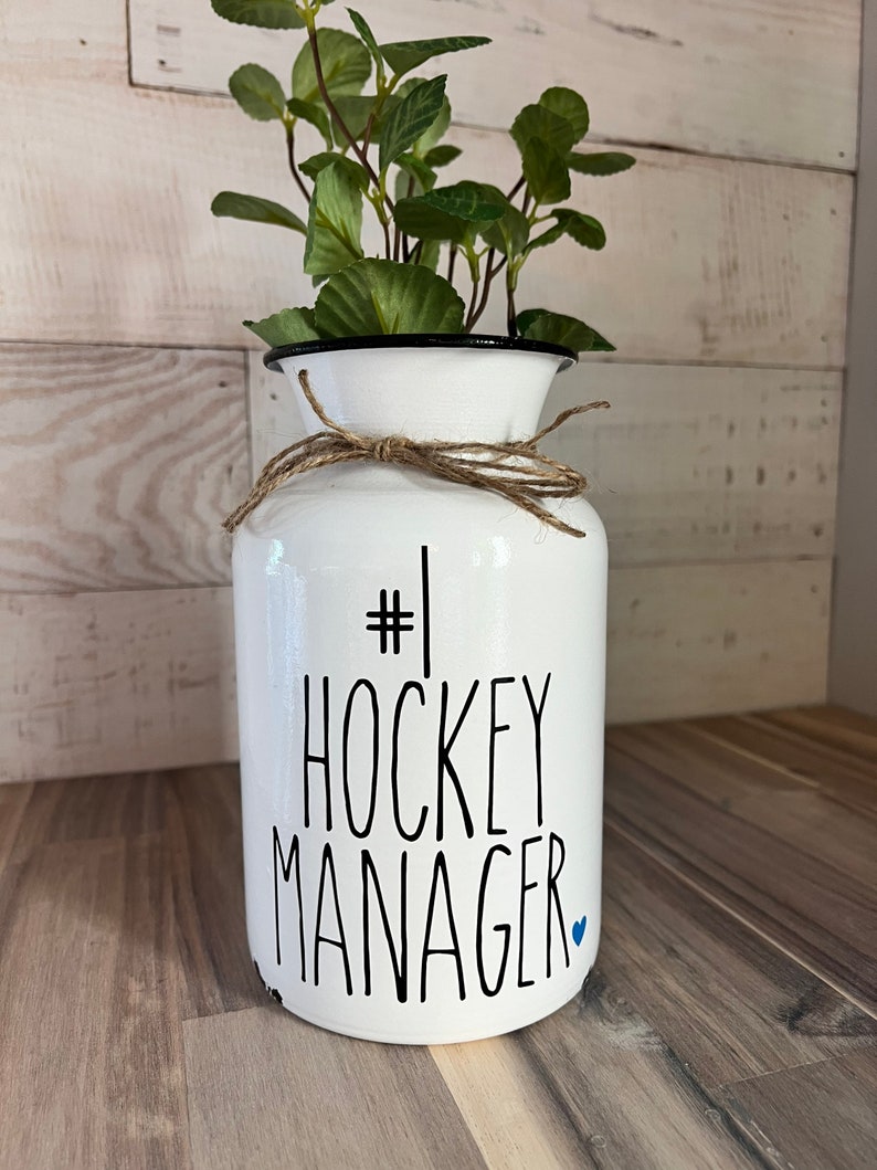 May include: White ceramic vase with a black rim and a brown twine bow. The vase has the text "#1 HOCKEY MANAGER" written in black with a small blue heart.
