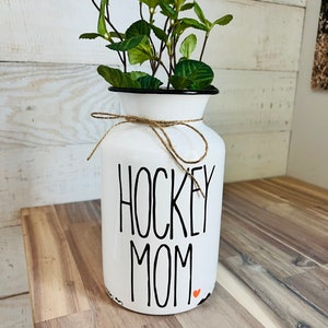 May include: White ceramic vase with a black rim and a brown twine bow. The vase has the words "HOCKEY MOM" written in black lettering with a small red heart.