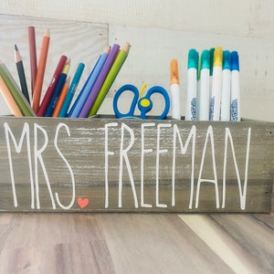 May include: A gray wooden box with the words "MRS FREEMAN" written in white paint. The box has a small red heart painted on the front. The box is filled with colored pencils, markers, and scissors.