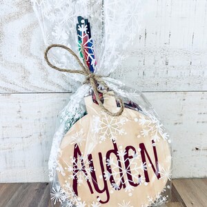 Może przedstawiać: Drewniana ozdoba świąteczna z personalizowanym napisem "Ayden" w czerwonych literach, otoczona białymi płatkami śniegu. Ozdoba jest zapakowana w przezroczysty plastikowy woreczek z białym wzorem płatk&oacute;w śniegu i związana sznurkiem.