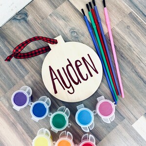 Może przedstawiać: Drewniana ozdoba z namalowanym imieniem "Ayden", wraz z zestawem słoiczk&oacute;w z farbą i pędzlami. Ozdoba ma czerwoną i czarną wstążkę.