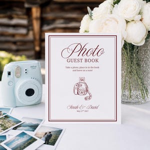 Può includere: Un cartello bianco con bordo rosso scuro e il testo Photo Guest Book, Take a photo, place in in the book and leave us a note!, Sarah & David, May 27th 2027, con un disegno al tratto di una fotocamera istantanea accanto a una fotocamera azzurra.