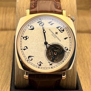 May include: A gold-tone wristwatch with a textured cream dial, black Arabic numerals, and a brown alligator-pattern leather strap. The watch face features the text Vacheron Constantin Geneve and a visible tourbillon movement at the six o'clock position.