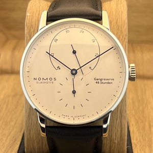 May include: A Nomos Glashütte wristwatch with a cream-colored dial, black hands, and a black leather strap. The dial features a power reserve indicator with the text Gangreserve 48 Stunden, a small seconds sub-dial, and the text Made in Germany.