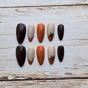 May include: A set of ten almond-shaped press-on nails featuring a glossy finish in shades of dark brown, cream, and burnt orange. Some nails are solid colors, while others feature a cream base with a hand-painted orange pumpkin design.
