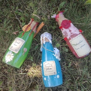 Może przedstawiać: Trzy ozdobne szklane butelki z etykietami Forest Dew, Mermaid Potion i Aphrodite's Love Potion. Zielona butelka ma stokrotkę, niebieska ma białe perły, a różowa jest ozdobiona koronką, czerwonymi pieczęciami lakowymi i małymi czerwonymi kwiatami.