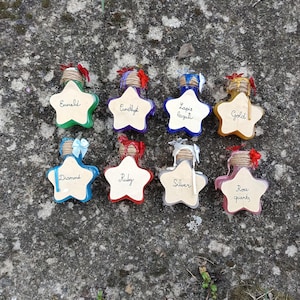 Könnte beinhalten: Acht sternförmige Glasflaschen mit farbiger Flüssigkeit, jede mit einem mit Schnur umwickelten Hals, einer kleinen Schleife und einem beigen sternförmigen Etikett. Die Etiketten sind mit den Namen Emerald, Amethyst, Lapis lazuli, Gold, Diamond, Ruby, Silver und Rose quartz beschriftet.