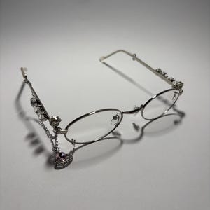 Peut inclure: Lunettes ovales argentées avec des verres transparents et une fine monture en métal. Les branches sont ornées de petits cristaux transparents et une fine chaîne argentée pend sur le côté gauche, terminée par un pendentif en forme de cœur rose.