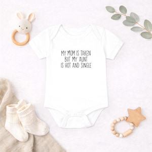 Puede incluir: Un body blanco de manga corta para bebé con el texto impreso en negro MY MOM IS TAKEN BUT MY AUNT IS HOT AND SINGLE. La prenda tiene cuello redondo y cierre de botones a presión en la parte inferior, junto a un sonajero de conejo y calcetines.