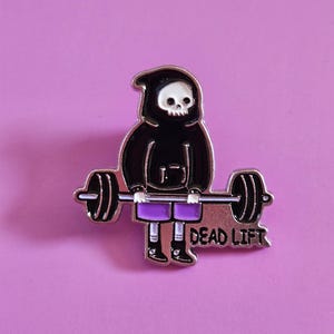 May include: A metal enamel pin featuring a cartoon skeleton wearing a black hooded sweatshirt and purple shorts, performing a deadlift with a barbell. The text "DEAD LIFT" is printed in black capital letters on the right side of the pin.