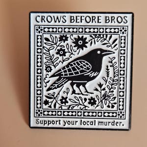 May include: A rectangular enamel pin featuring a black crow surrounded by white floral patterns within a geometric border. The text CROWS BEFORE BROS is printed at the top, and the text Support your local murder. is printed at the bottom.