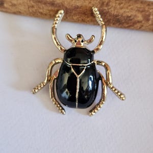 Op de afbeelding: Een goudkleurige metalen keverbroche met een glanzend zwart geëmailleerd lichaam. Het insectontwerp heeft gesegmenteerde poten en gebogen antennes, met een gladde, reflecterende afwerking op het centrale schild en een gouden metalen rand.
