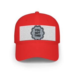 Könnte beinhalten: Eine leuchtend rote Baseballkappe mit einem horizontalen, weißen, rechteckigen Aufnäher auf der Vorderseite. In der Mitte des Aufnähers befindet sich ein graues Emblem mit gewelltem Rand und dem schwarzen Sticktext "YOU GOT THIS!".