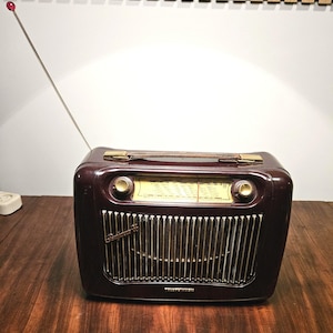 Puede incluir: Una radio portátil de color granate oscuro con un dial de frecuencia crema, dos perillas doradas y una rejilla vertical plateada. La radio cuenta con un asa de cuero marrón, una antena larga extensible con punta roja y el texto Telefunken.