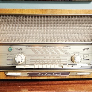 Op de afbeelding: Een vintage Graetz Melodia radio met een gepolijste houten behuizing, een metalen luidsprekerrooster en een centraal bedieningspaneel met twee grote ronde knoppen, diverse drukknoppen en gedrukte frequentieschalen in zwarte tekst op een zilveren achtergrond.
