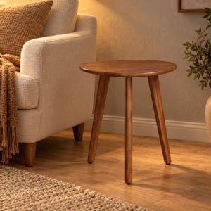 May include: A round wooden side table with a smooth, medium-brown finish, featuring three tapered legs that extend outward from the center. The table has a simple, circular top surface and a sturdy construction made of natural wood grain.