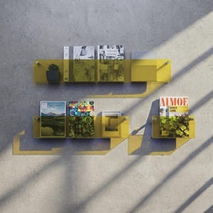 May include: Two horizontal rows of yellow translucent acrylic wall shelves mounted with silver hardware. The shelves hold various books, a small potted plant, a framed picture, and a magazine with the text AIMOE SHNEE SHNE on the cover.