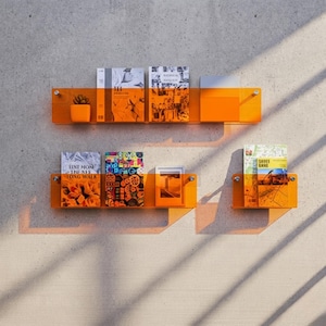 May include: Two horizontal orange acrylic wall shelves mounted on a wall. The top shelf holds a small orange planter and three books. The bottom shelf holds two books, a small framed picture, and a single book on a separate smaller shelf section.