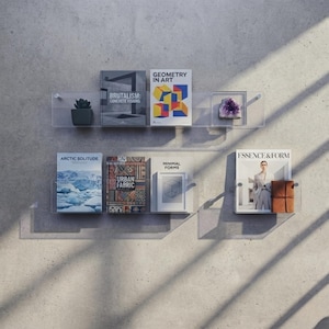 Peut inclure: Deux étagères murales flottantes en acrylique transparent contenant divers livres et objets décoratifs. L'étagère supérieure présente une petite plante en pot, des livres intitulés Brutalism: Concrete Visions et Geometry in Art, et un cristal violet. L'étagère inférieure contient des livres intitulés Arctic Solitude, Urban Fabric, Minimal Forms et Essence & Form.