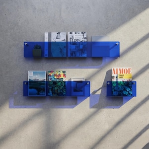 May include: Three wall-mounted shelves made of translucent blue acrylic material. The shelves are arranged in two rows and hold various books, a small potted plant, and a framed picture. The shelves are secured with visible silver metal hardware.
