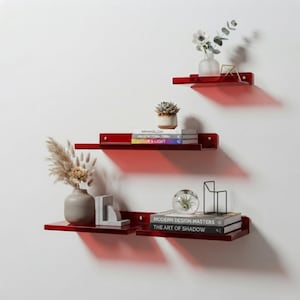 Peut inclure: Trois étagères flottantes rouge brillant fixées au mur. L'étagère supérieure porte une fleur blanche dans un vase. L'étagère du milieu présente une petite plante grasse et trois livres intitulés Minimalism, Architectural Form et Color & Light. L'étagère inférieure contient un vase avec des plantes séchées, un objet géométrique gris, un globe en verre avec une plante, un cadre en fil métallique et deux livres intitulés Modern Design Masters et The Art of Shadow.